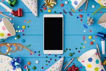Top view photo of birthday composition party accessories and smartphone with copyspace in the middle on isolated blue wooden table background