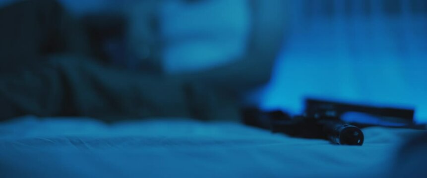 Close up of M16 rifle lying on a bed, with PTSD soldier siiting on bed in the background.