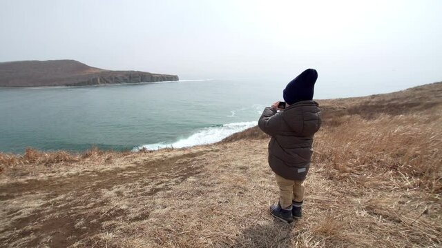 Back of a small boy taking photo of beautiful marine landscape: blue sea and high shores. Spring.