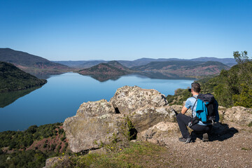 Un randonneur de dos , accroupis en train de prendre une photo  d'un lac bleu et des montagnes qui l'entourent