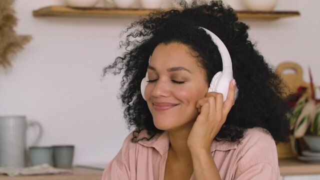 African American woman in big white headphones listens to music with pleasure using smartphone. Black brunette woman posing sitting at table bright kitchen. Close up. Slow motion ready, 4K at 59.97fps