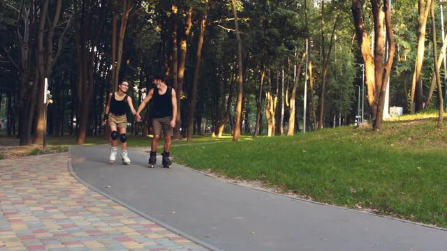 Young happy couple rollerblading in the park holding hands. Young attractive girl and a young guy rollerblading in the park. Young couple in love enjoy roller skating in a summer park.