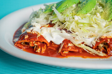 Exquisite traditional Mexican red enchiladas with chopped lettuce, sour cream, cheese and avocado on a white plate.