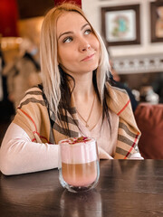 A blonde girl with a scarf on her shoulders in a soft pink sweater sits in a cozy restaurant and she is relaxed and have a coffee drink and a piece of cake on her table.