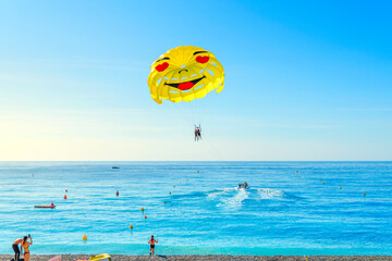 Tourists in bathing suits watch as a boat with a parasailing couple takes off over the Bay of Angels on a sunny summer day on the French Riviera