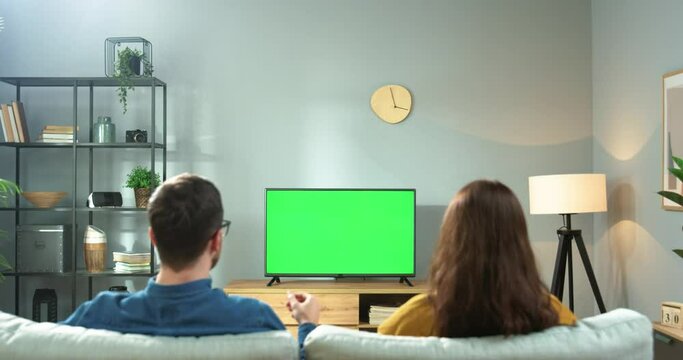 Rear of young Caucasian happy couple man and woman sitting on couch in living room watching at TV monitor with green screen having fun. Family cheering for fav team, watching TV, sport match