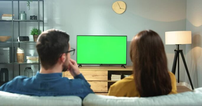 Back view of Caucasian couple wife and husband watching TV with chroma key and eating snacks enjoying sport game Man and woman cheering for fav team raising hands watching football match on television