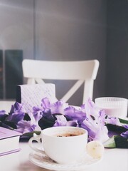 Composition with a cup of coffee, a book, violet Gladiolus flowers bouquet, a candle and a map print on a white table top with a chair on a background.