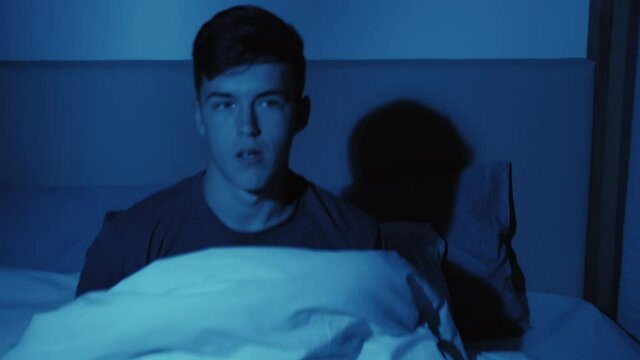Night terror. Sleep disorder. Bedtime mystery. Panic attack. Disturbed paranoid guy raised up in bed scared by nightmare of monster hand shadow on wall late at home in dark blue light.