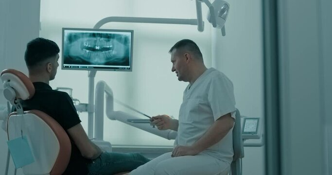 A male patient is sitting in the dentist chair and looking at the monitor screen with human jaw x-ray while a male dentist explaining something and using pointer to show a teeth.