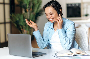 Satisfied successful mixed race stylish business lady, manager, freelance, wearing headset, working remotely, communicates by online call with a colleague or client, gesturing with her hand, smiling