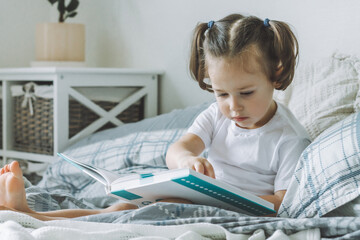 Little dark-haired girl 2-4 with two ponytails sits on bed with pillows and reads book