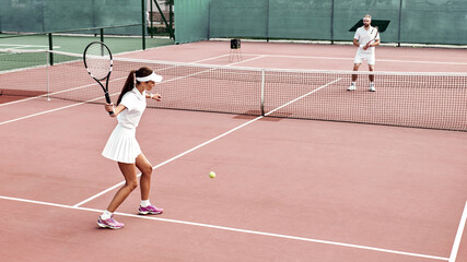 Pretty girl plays tennis on the court outdoors. She prepares to beat on a ball. Woman wears a light blue sportswear with white sneakers