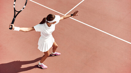 Pretty girl plays tennis on the court outdoors. She prepares to beat on a ball. Woman wears a light blue sportswear with white sneakers. Top view photo.