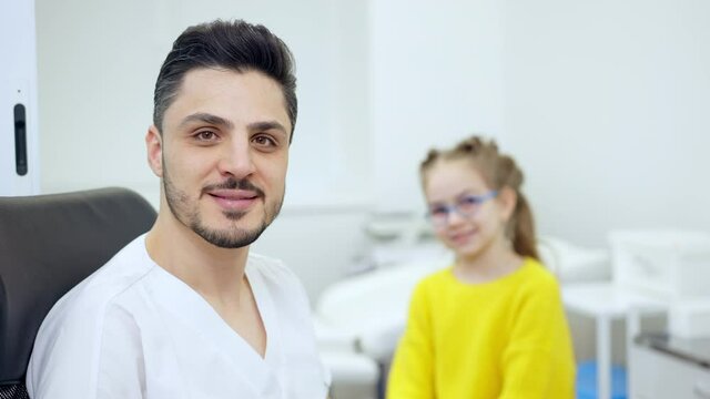 Portrait of confident male Middle Eastern pediatrician posing in hospital with blurred Caucasian little girl at background. Positive handsome man looking at camera smiling consulting patient.