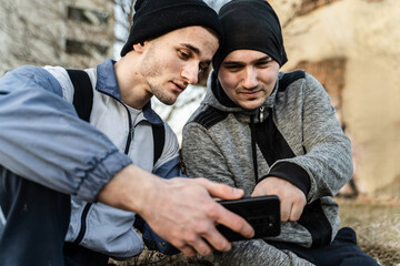 Portrait of two adult men male migrants sitting outdoor in rural environment in cold day checking location on mobile phone using navigation gps app on maps to plan the route for travel - obrazy, fototapety, plakaty
