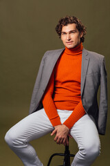 Tall handsome man dressed in orange turtleneck, grey jacket and white jeans posing on the green background