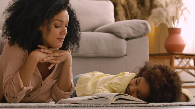 Afro American woman reads book with an interesting fairy tale to little girl. Mother and daughter posing, lying on floor in room with light home interior. Close up. Slow motion ready, 4K at 59.97 fps.