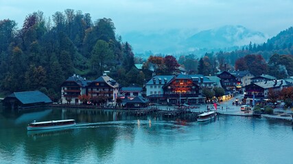 Obraz premium Sightseeing boats cruising on Konigssee ( King's Lake ) with lakeside hotels, resorts and boathouses at foggy misty dusk ~ Beautiful fall scenery of Bavarian countryside in Berchtesgaden Germany