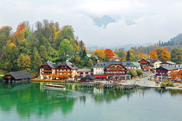 Fototapeta na wymiar A sightseeing boat cruising on Konigssee ( King's Lake ) surrounded by colorful autumn trees and boathouses on a foggy misty morning~ Beautiful scenery of Bavarian countryside in Berchtesgaden Germany
