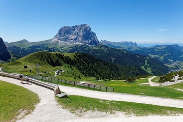 Summer scenery of rocky Mountains Sassolungo (Langkofel) & Schlern (Sciliar) with view of a cable car at mountainside & tourists on hiking trails in the valley in Selva di Val Gardena, Dolomiti, Italy