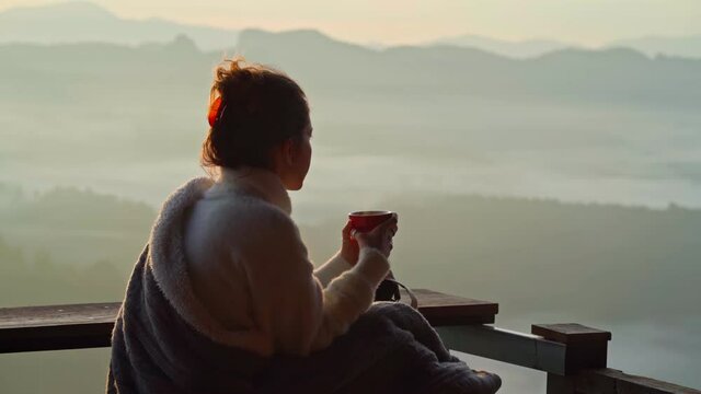 young woman is sitting with a mug of coffee on a cliff overlooking the mountains with fog in the morning