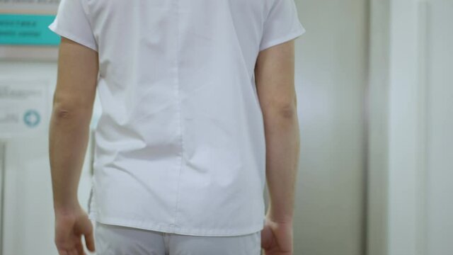 Back view of young unrecognizable man in medical uniform walking along corridor to hospital lift. Camera follows Middle Eastern doctor in clinic indoors.