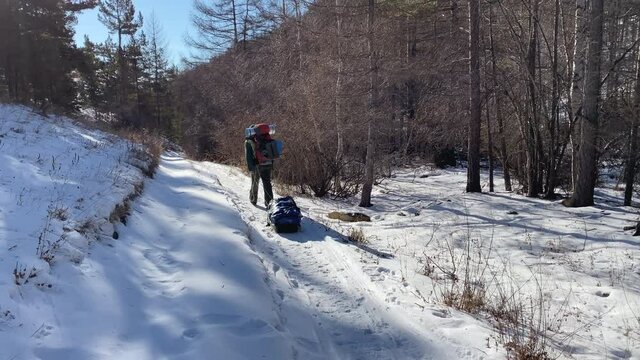 A guy with a backpack goes on a winter hike and drags along with him a sack with a large bag of supplies and equipment. Beautiful winter landscape. Olkhon Island. Snow-covered trail in the forest.