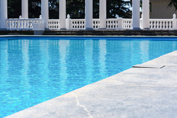 Swimming pool with clear blue water in the garden. Decorative decoration of the park. A clear, warm summer day. White columns, tall trees, a landscape without people. The concept of summer holidays.