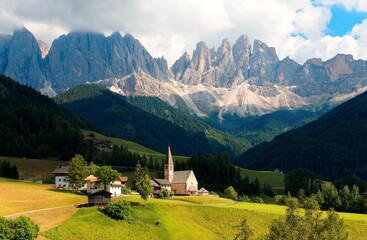 Idyllic scenery of Val di Funes on a cloudy summer day with rugged peaks of Odle mountain in background & a church in Village Santa Maddalena in the green grassy valley in Dolomiti, South Tyrol, Italy
