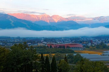 Fototapeta na wymiar Sunrise scenery of majestic Hakuba Mountain Range with golden glow on the submit, a fog belt on the mountainside and village houses in the field on a foggy autumn morning in Nagano, Japan