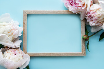 Wooden frame surrounded by beautiful pink peonies on a blue background, top view, copy space, flat lay.