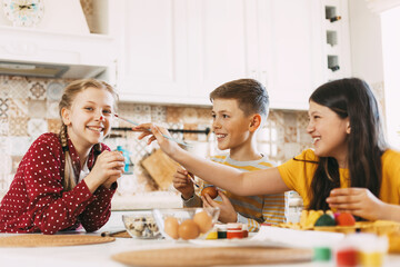 Two sisters and a brother are sitting at the table and painting Easter eggs in different colors for Easter, laughing and indulging