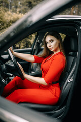 Naklejka premium A beautiful young girl in a red overalls sits behind the wheel of a black car on an empty road in the forest