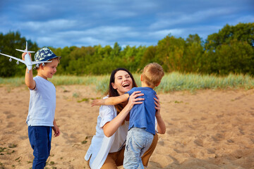 A young happy mother with two boys playing on the sand near the forest, in the hands of one of them a model of a civil aircraft and a hat on his head
