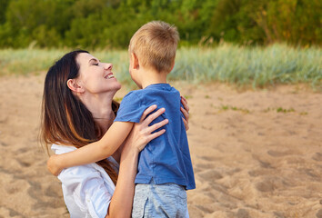 Mother with long hair and a smile turned to her son and hugs him against the background of the forest and sand