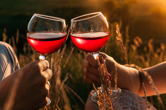 Two hands toasting red wine glasses. Toast and clinking glasses. Romantic dinner for a couple in love. Outdoor picnic in the grass in summer at sunset. the rays of the sun are reflected in the glasses