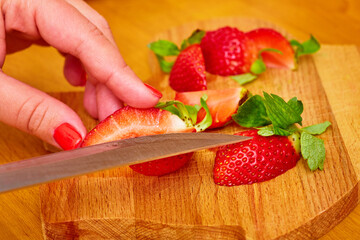 On the cutting Board are several ripe juicy strawberries, one of them is cut by a steel knife in a woman's hands