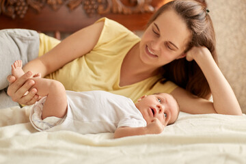 Mother and baby lying on bed on white blanket, smiling mommy wearing yellow t shirt enjoying to spend time with her newborn kid, infant looking away to study outward things.