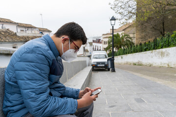 Teenager wearing glasses and protective face mask using his mobile phone while sitting in the street.