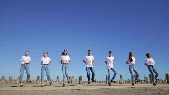 A group of young people in white T-shirts are dancing a tap outdoors, against a background of blue sky.