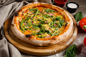 Homemade pizza with arugula, pickles, ham and sauce on a wooden pizza plate.