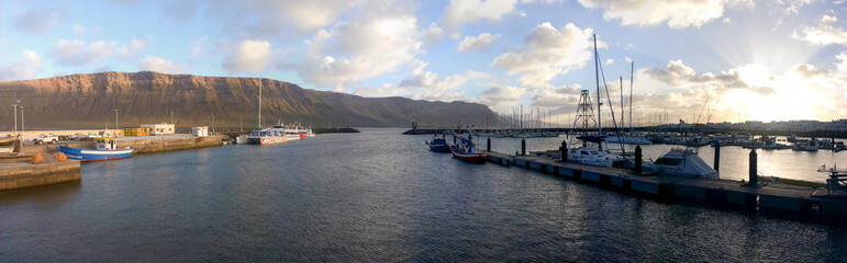 Fototapeta premium Views from Caleta de Sebo at dusk, with the fishing and tourism boats docked at the main pier on the island of La Graciosa and with the cliff of Famara, on the island of Lanzarote, in the background.
