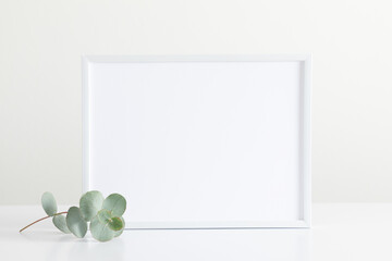White frame mockup, green eucalyptus leaves on white table. Front view. Place for text, copy space,...