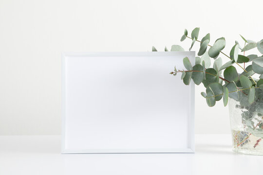 White frame mockup, green eucalyptus leaves on white table. Front view. Place for text, copy space, mockup