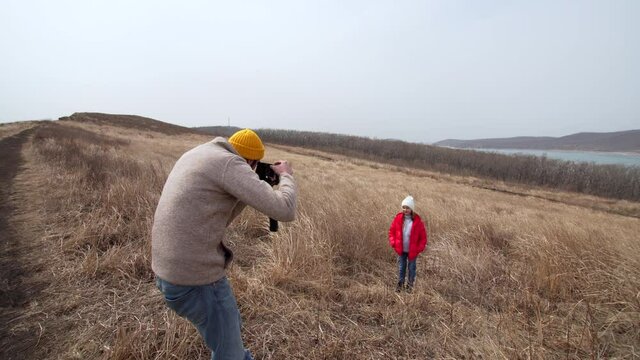 Father is taking photos of his cute daughter in dry grass. Sea bay is on the background. Early spring.