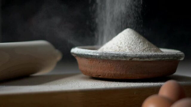 Flour being sieved into a rustic bowl. Baking ingredients and equipment included in frame. Muted colour palette. Slow motion falling flour against a dark background.