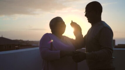 Happy Latin senior couple having romantic moment dancing on rooftop during sunset time - Powered by Adobe