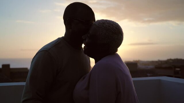 Happy Latin senior couple having romantic moment kissing on rooftop during sunset time