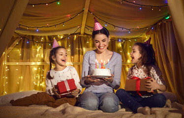 Children make wonderful birthday surprise gift for their mom. Happy young mother getting presents and cake with candles from her cute little twin daughters at fun party at home